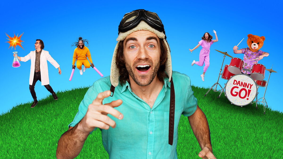 Danny GO! in an aviator hat smiling on grass under blue sky, with scientist holding beaker, person with pink bats, woman jumping, and bear costume playing drums labeled "Danny Go!" in colorful, playful setting.