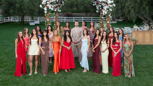 A group of women in colorful dresses and one man in a beige suit stand outdoors on grass, under floral arches, surrounded by trees and wooden fences, in a formal garden setting.