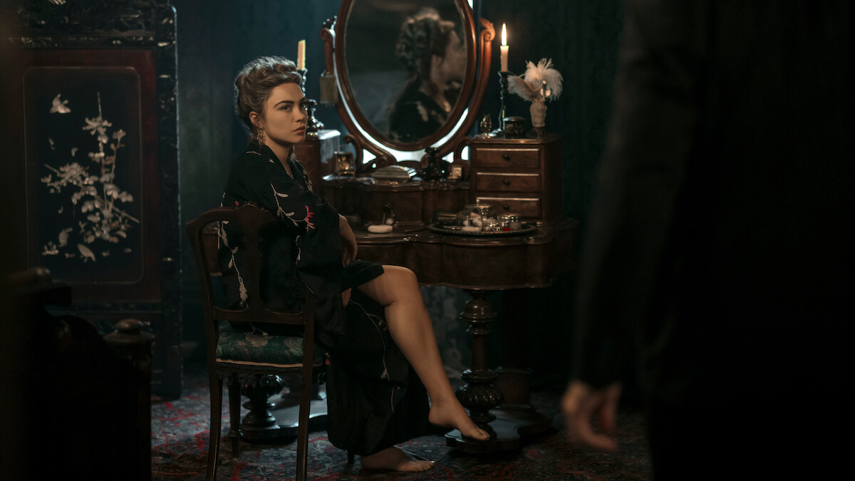 Woman in a dark robe sits at a vintage vanity with candles and flowers in a dim, elegant room, looking toward a standing figure partially visible in the foreground.