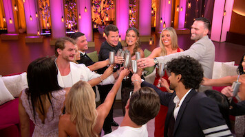 A group of people standing in a circle raising metal cups in a celebratory toast, dressed formally, in a brightly lit room with purple and pink lighting, lounge seating, and decorative hanging lights in the background.
