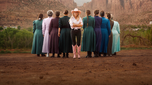 A group of women in long, modest dresses stand in a dirt area, backs to the camera, with one woman in a cowboy hat, white shirt, black pants, and pink boots facing forward against a backdrop of mountains and greenery.