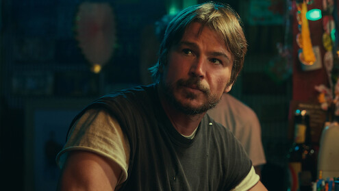 Man with short brown hair and beard sits at a dimly-lit bar, wearing a dark T-shirt over a light shirt. Bar items and ambient lighting are visible in the colorful, casual setting.