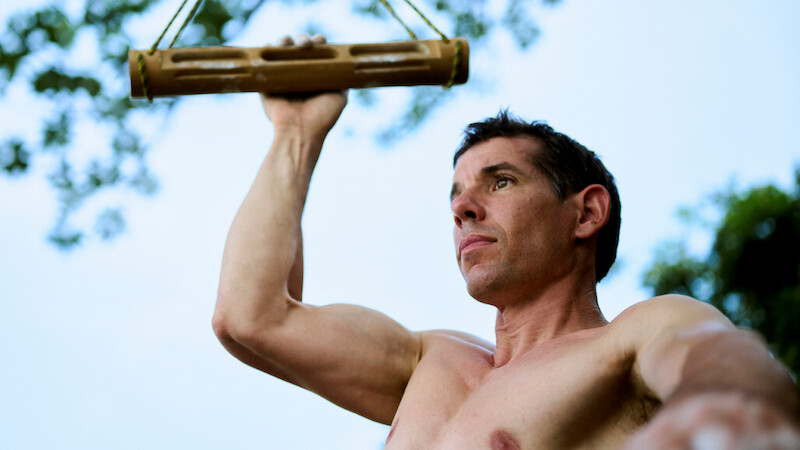 Alex Honnold outdoors using a wooden grip training tool, surrounded by trees and sky, focused and exercising for strength or climbing fitness.