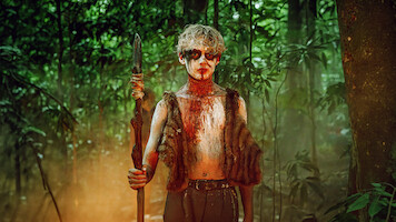 A person with painted face and fur vest stands holding a spear in a dense, misty, green forest, suggesting a primitive or survival setting.