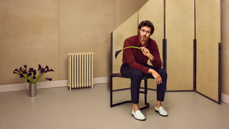 Adam Brody está sentado em um banco segurando uma flor copo-de-leite preta