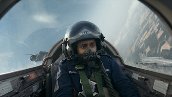 Pilot in cockpit flying a jet fighter, wearing helmet and oxygen mask, with intense expression; another jet visible outside, sky and landscape blurred from high speed and aerial maneuver.