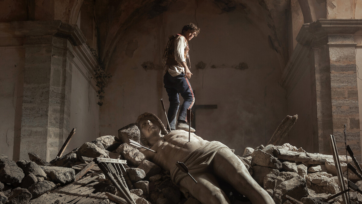 A person stands on rubble inside a damaged, old stone building, with a large broken statue lying among the debris, creating a dramatic and somber atmosphere.