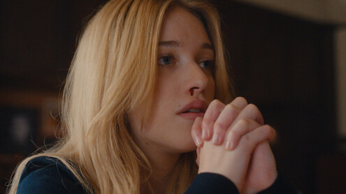 Young woman with long blonde hair sitting indoors, resting her chin on clasped hands, looking thoughtful or concerned, with a blurred background and warm lighting.