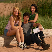 Three young adults sitting on a log at a sandy outdoor area with tall grass, dressed in casual summer clothes; one holding papers, all looking at the camera and smiling, suggesting a relaxed, sunny environment.