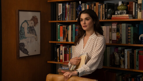 Rachel Weisz as The Protagonist  in a white blouse stands in front of a bookshelf filled with books and decor, holding folders. A framed artwork hangs on the wall, creating a cozy, intellectual office or library setting.