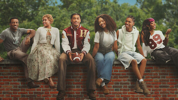 Six young adults sit on a red brick wall outdoors at Hillman College, dressed in casual and vintage clothing, smiling and relaxed, with green trees in the background.