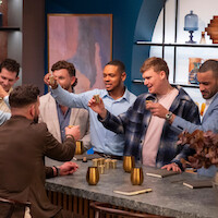 Group of men socializing around a modern bar counter, raising gold cups in a celebratory toast. The setting features stylish decor, notebooks, and warm lighting, creating a casual, contemporary atmosphere.