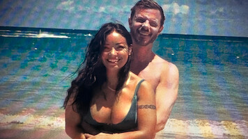 A smiling couple standing together on a sunny beach with waves and a blue ocean in the background.