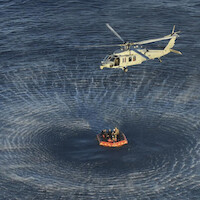 Helicopter hovering above ocean, creating circular ripples, while a group of people wait in an orange life raft below, possibly during a rescue or training operation at sea.