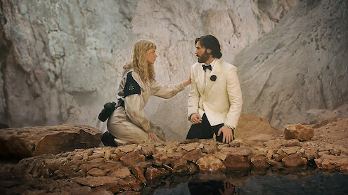 A woman in an adventure outfit kneels by a man in a white tuxedo; they appear to be talking in a rocky, cave-like environment near a shallow pool of water.