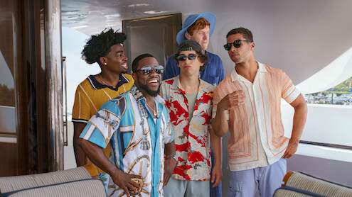 Five men in colorful summer shirts are standing and talking on a luxury boat deck, smiling and laughing together in a relaxed, sunny outdoor setting.