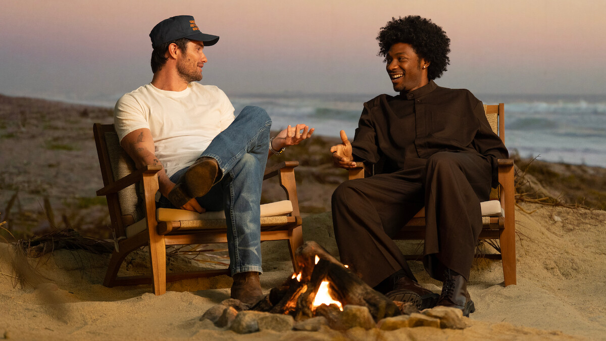 Two men sitting in chairs on a sandy beach at sunset, talking and smiling by a small campfire with the ocean and waves in the background.