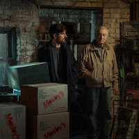 Three people stand in a dimly lit, cluttered basement or storage room with brick walls and stacked boxes. The mood appears tense or serious, and the setting looks old and industrial.