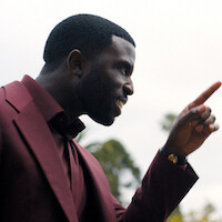 Man in a burgundy suit pointing and speaking with intensity outdoors, with blurred trees and cloudy sky in the background.