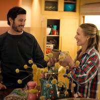 Two people smiling and exchanging mugs in a cozy kitchen, surrounded by yellow cabinets, flowers, and assorted kitchen items, suggesting a warm and casual atmosphere.