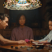 Three people sit quietly around a wooden table under a colorful stained-glass lamp in a dimly lit room; a man to the left, a woman in the center, and a child on the right focused on drawing.