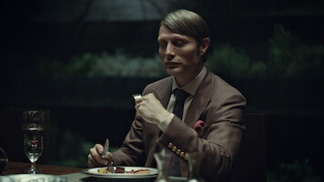 A well-dressed man sits alone at a dimly lit dinner table with a formal place setting, holding a fork and knife, ready to eat. The background is dark and blurred, suggesting an elegant, intimate indoor setting.