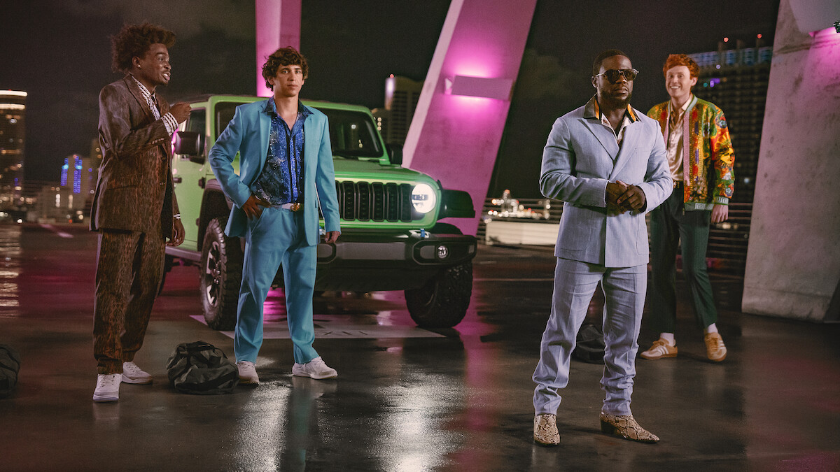Four men in stylish suits stand in front of a green Jeep on a rooftop at night, illuminated by pink and purple lights, with a cityscape in the background.