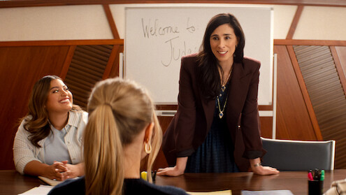 Three women in a meeting room, one standing and smiling while leaning on a table, two seated, with a whiteboard in the background that says "Welcome to". Professional office setting.