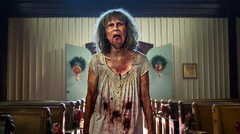 A disheveled, bloodied woman with gray hair stands in the aisle of a decorated church, with pews on either side and wreaths on the doors in the background, looking distressed or possibly zombified.