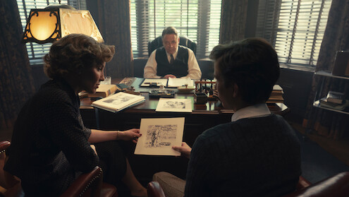Two people sit across from a man at a desk in a dimly lit office, discussing a drawing. The office has large windows with blinds, books, and papers scattered on the desk, creating a serious, professional atmosphere.