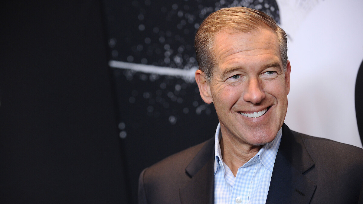 Man in a suit smiling in front of a dark, abstract background with white speckles and lines, possibly at an event or exhibition.