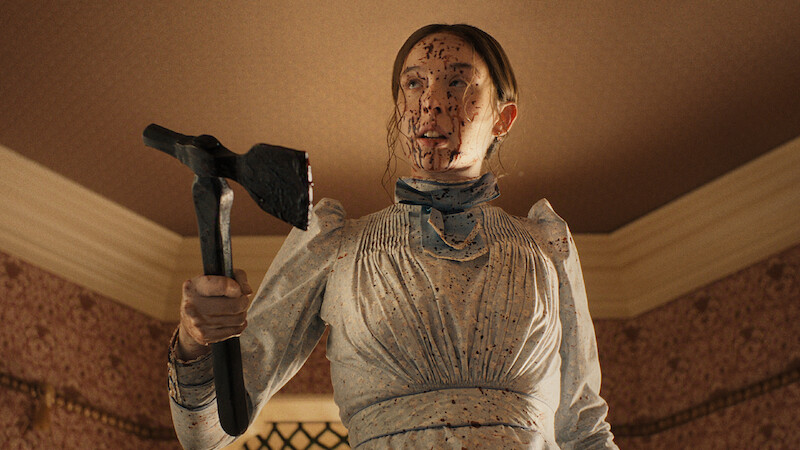 Woman in an old-fashioned white dress, splattered with blood, holding an axe inside a Victorian-style room with patterned wallpaper and a high ceiling, looking intense and determined.