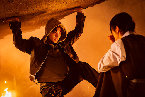 A person in a hoodie holds up a collapsing ceiling in a smoky, fire-lit room while another person crouches nearby, looking at them in concern.