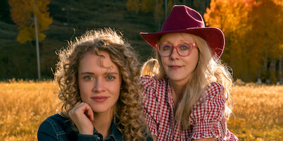Two women in a field with autumn trees; one with curly hair wearing denim, the other in glasses, a red hat, and checkered shirt. Warm sunlight, outdoors, fall setting.