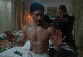 A man in a towel holds up his hands as a woman in uniform points a gun at the back of his head.