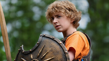 A young person with curly blonde hair in an orange shirt and armor holds a decorated shield outdoors, surrounded by green trees and natural light, appearing alert and determined.
