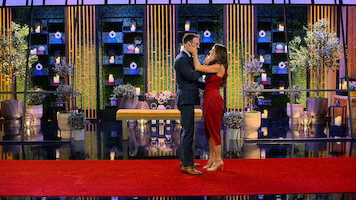A couple stands on a red carpet, embracing in a decorated, modern indoor setting with potted plants, candles, and geometric décor on shelves in the background. Elegant, romantic atmosphere with formal attire.