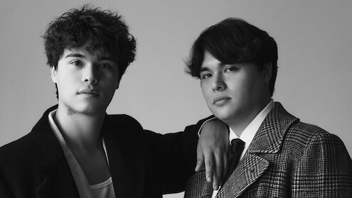 Two young men pose closely together in a studio setting, one resting his arm on the other's shoulder. Both wear jackets; the environment is minimal, and the photo is in black and white, giving a classic portrait feel.