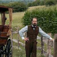 a man stands with a corn field on one side and a late 1800s carriage on the other. 