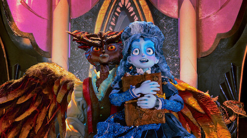 A blue-skinned, animated girl in a blue dress holds a book, standing nervously in front of a menacing owl-like creature with wings inside a lavish, ornately decorated room with pink and gold arches and dramatic lighting.