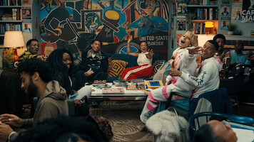 A diverse group of people laugh and socialize in a cozy, colorful living room filled with art, books, and a large mural, creating a lively, welcoming atmosphere.