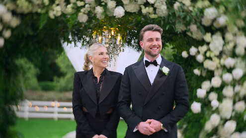A smiling man in a tuxedo and a woman in formal attire stand under a floral arch outdoors, surrounded by green foliage and white flowers, possibly at a wedding or formal event.