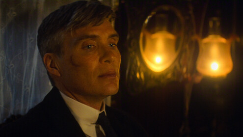 A man in a suit with a scar on his cheek sits in a dimly lit room, illuminated by warm yellow lamps and ornate décor, suggesting a vintage or historical setting.