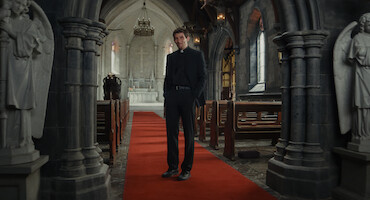 A priest stands on a red carpet in the middle aisle of a dimly lit stone church, flanked by statues, with pews on either side and an altar in the background, creating a solemn and contemplative mood.