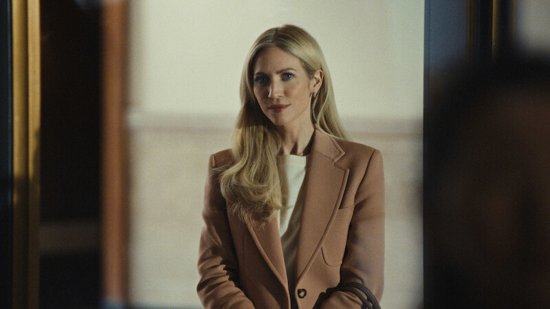 A woman with long blonde hair in a tan blazer stands indoors in a softly lit, neutral space, wearing a calm and confident expression, creating a professional and composed atmosphere.