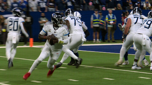 Football quarterback in white uniform running with the ball on a green field during a game, with teammates blocking and stadium spectators in the background.