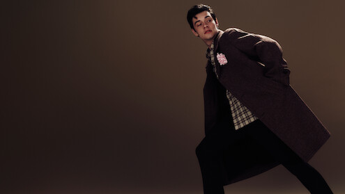 Louis Partridge stands on a dark brown background leaning over in an angled manner waring a brown coat with a plaid shirt and dark trousers.