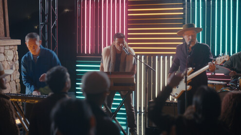 A band performs live on stage with colorful neon lights in the background, including a keyboard player, singer, and guitarist. Audience members are watching and raising their hands in an outdoor nighttime concert setting.