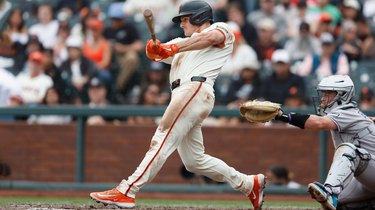 Matt Chapman in white uniform swinging bat at a pitch during a game, with catcher in gray behind him and a blurred crowd in stadium seats in the background.