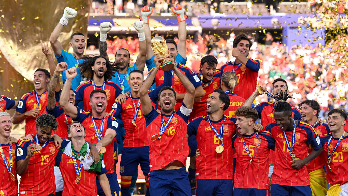 Soccer team in red and blue uniforms celebrates on a stadium stage, holding a gold trophy and wearing medals, with confetti falling and crowd in the background.